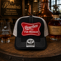 Trucker hat with 'Bourbon for Missiles' design on a wooden barrel with Blanton's whiskey bottles and glasses.
