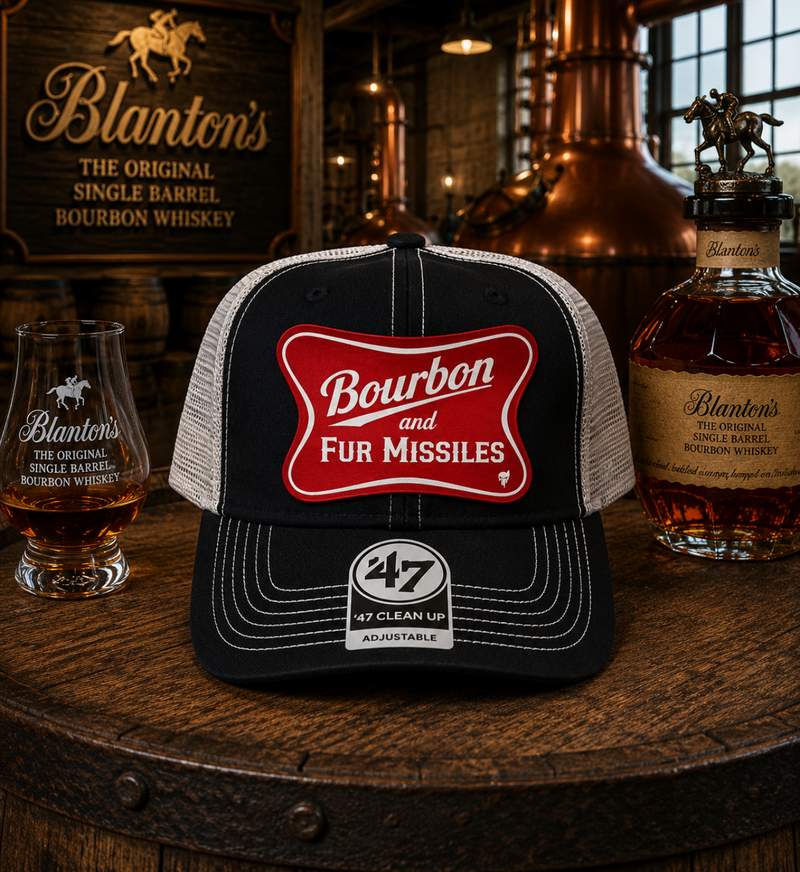 Trucker hat with 'Bourbon for Missiles' design on a wooden barrel with Blanton's whiskey bottles and glasses.